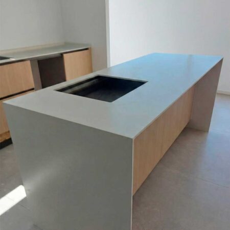 Isla de cocina moderna con encimera blanca, muebles de madera clara y hueco para fregadero o placa, con pared blanca de fondo.