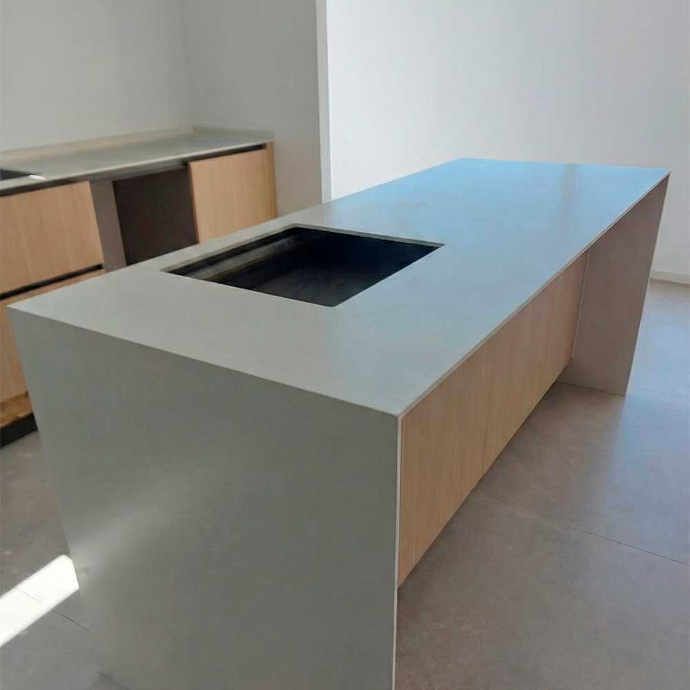 Isla de cocina moderna con encimera blanca, muebles de madera clara y hueco para fregadero o placa, con pared blanca de fondo.