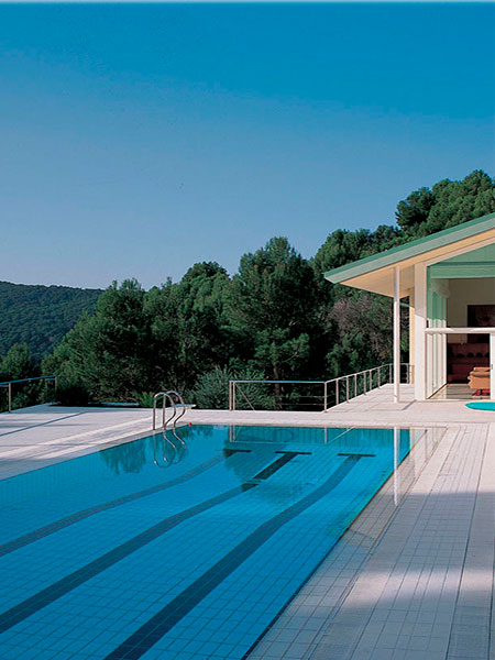 Piscina moderna revestida con cerámica blanca junto a vivienda de diseño rodeada de naturaleza.